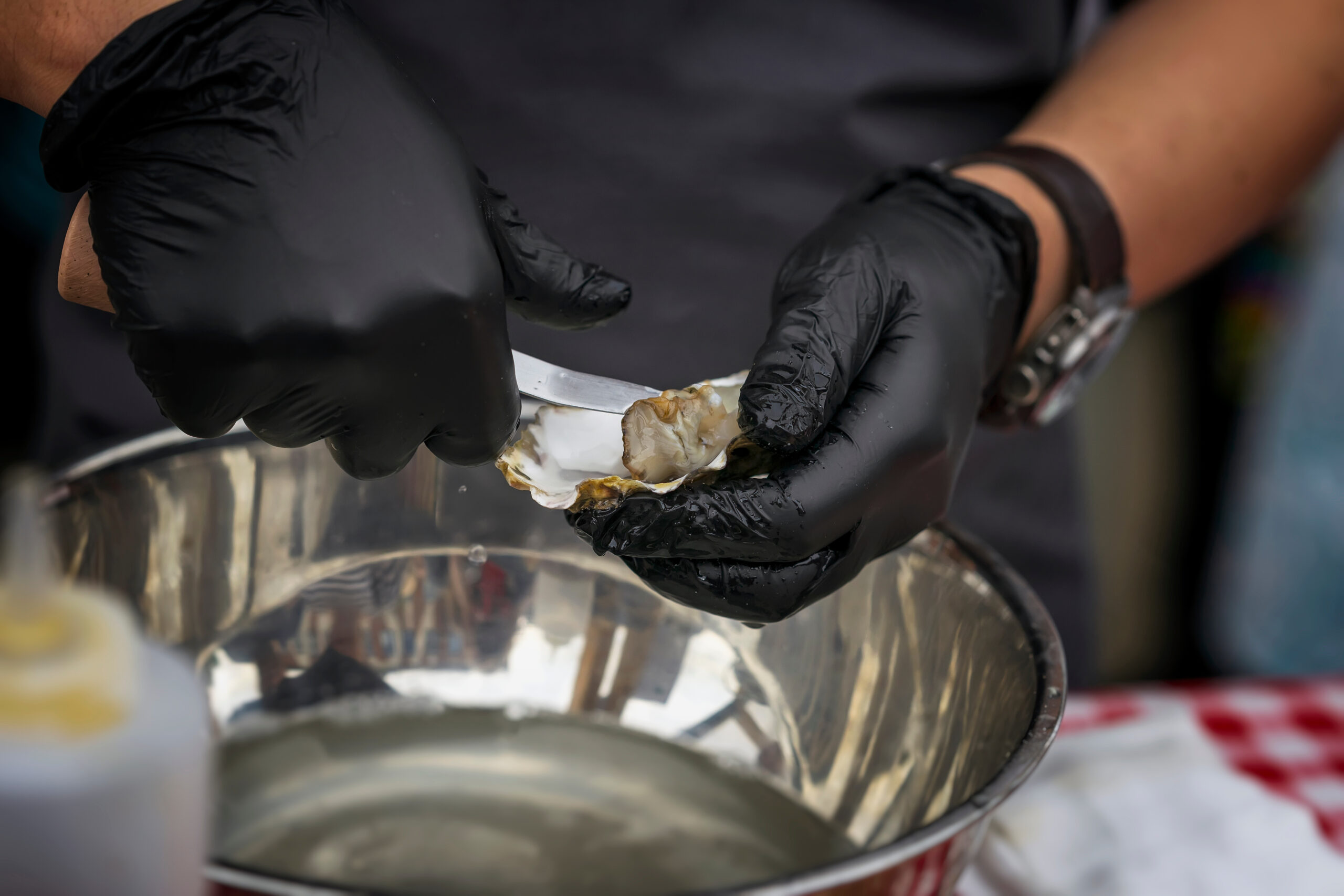 Shucking fresh oysters on a seafood market. Man’s hands in black gloves with knife. Gastronomic gourmet dainty products on market counter, real scene in food market